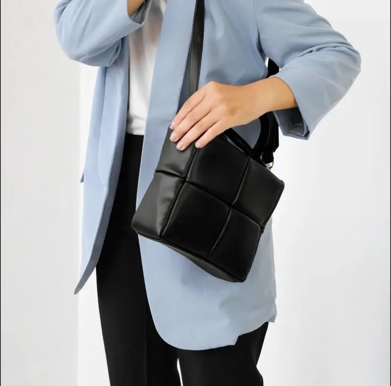 Wanita berblazer biru muda dan celana hitam memakai tas tangan hitam berpanel kotak dengan tali panjang di bahu, tangannya memegang bagian atas tas