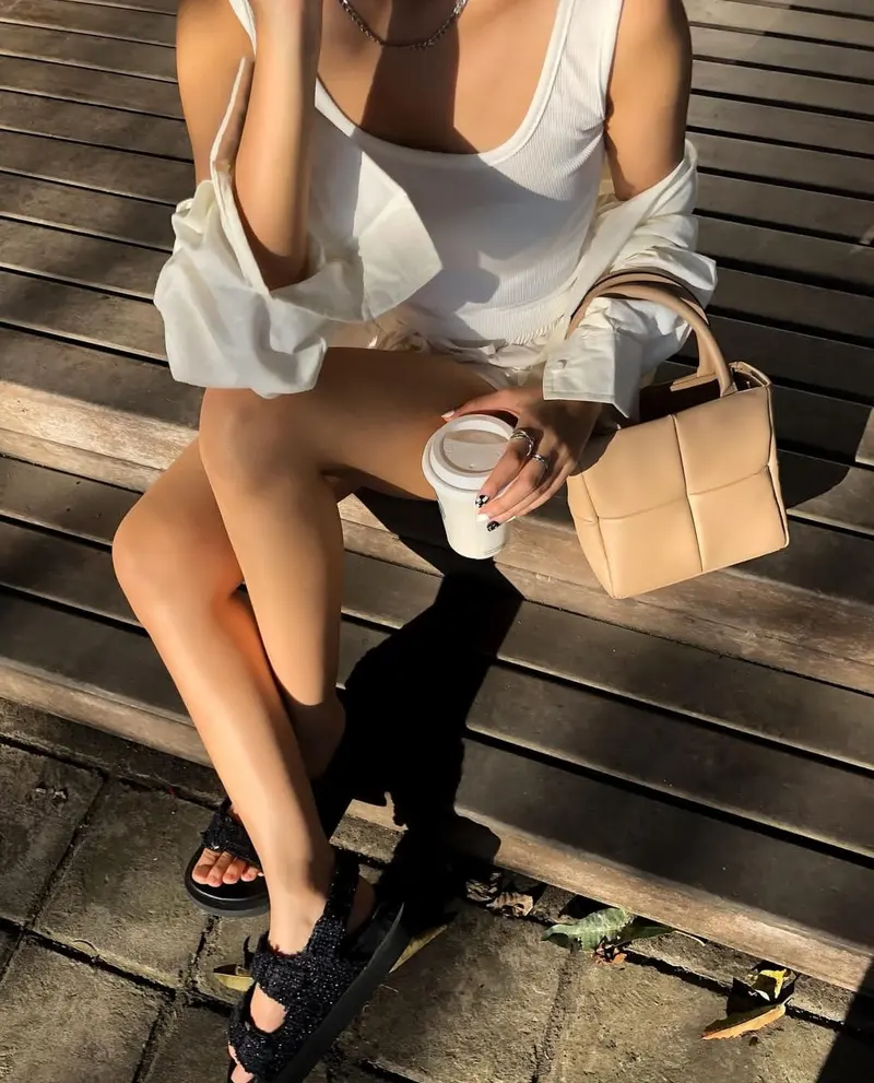 Wanita berkaus tanpa lengan putih dan kemeja longgar duduk di bangku kayu memegang gelas kopi, dengan tas tangan krem berpanel kotak di sampingnya