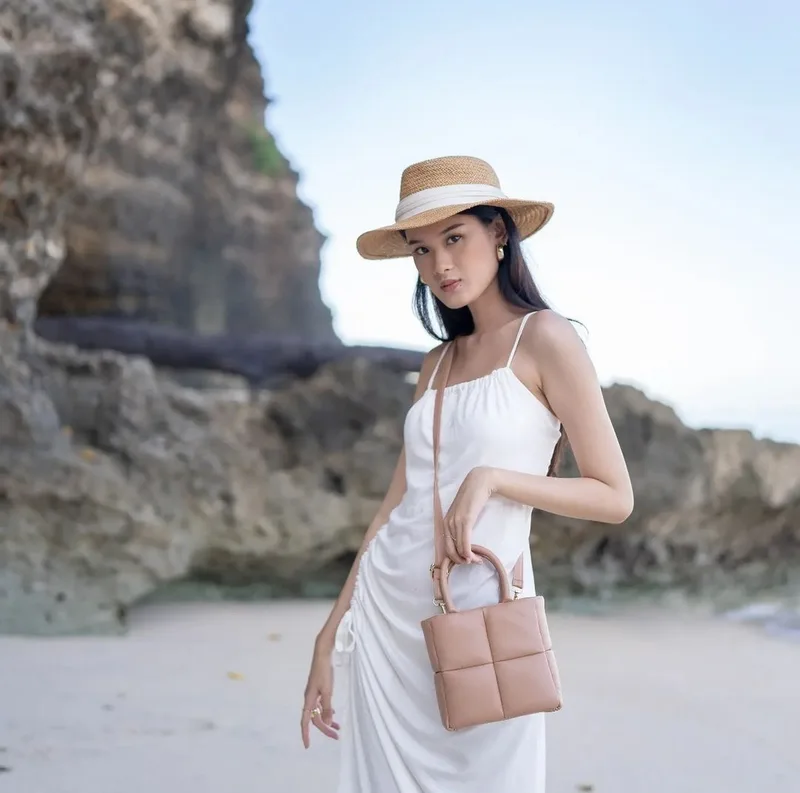 Wanita bertopi jerami dan bergaun putih bertali tipis di pantai berkarang memegang pegangan tas tangan nude berpanel kotak, tali panjang tergantung di bahu