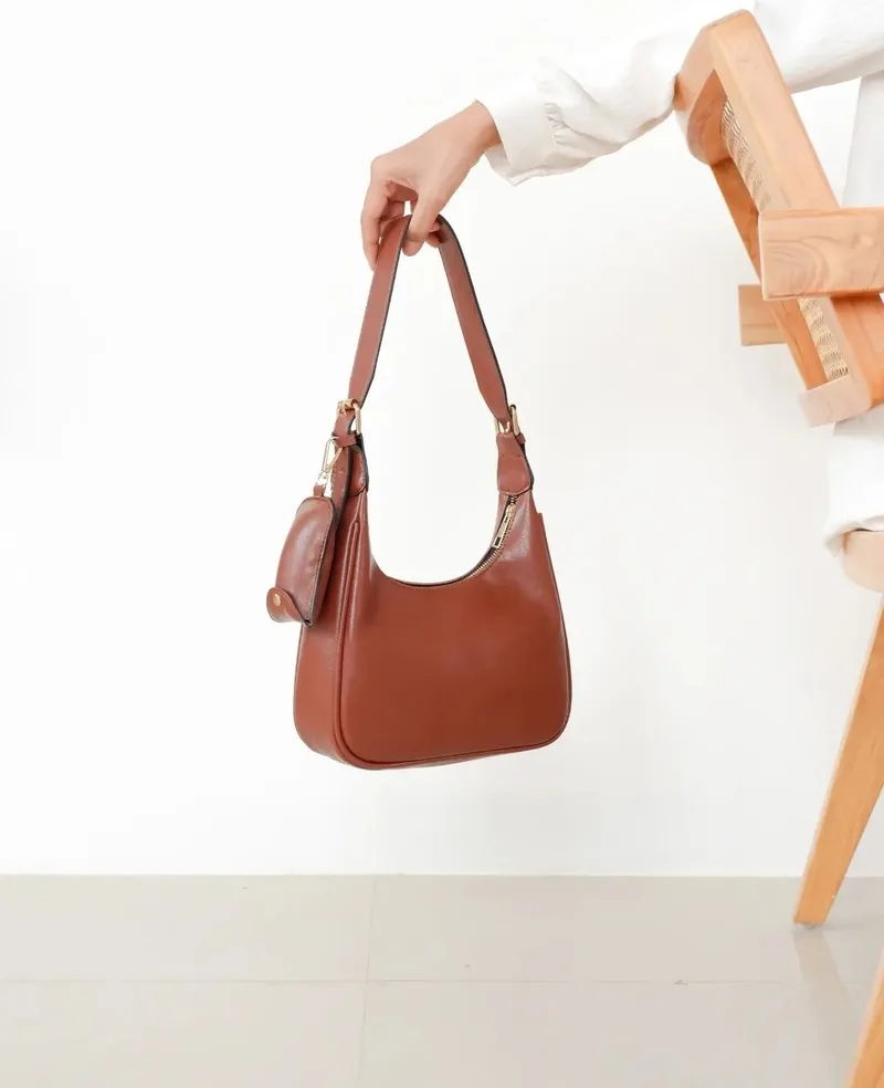 Tangan memegang tali tas bahu kulit sintetis cokelat bersiluet lengkung dengan pouch kecil tergantung di sisinya, di depan dinding putih dan kursi kayu
