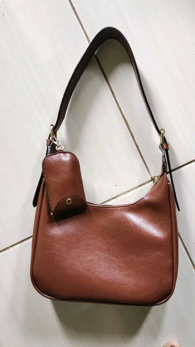 Tas bahu cokelat berbentuk setengah bulan dengan tali bahu lebar dan dompet kecil yang menggantung di samping, digantung pada dinding putih