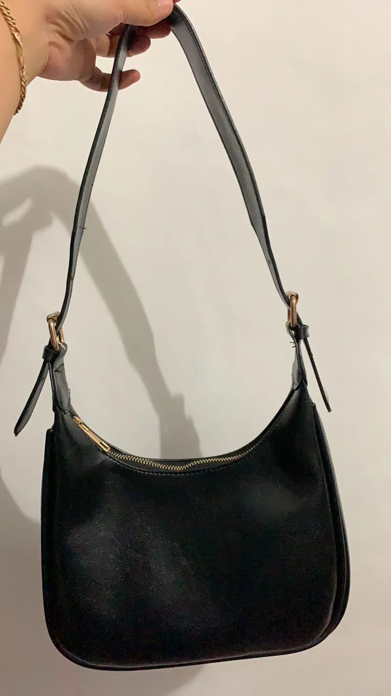 Tangan memegang tali tas bahu hitam berbentuk setengah bulan dengan gesper keemasan di kedua sisi tali, di depan dinding putih
