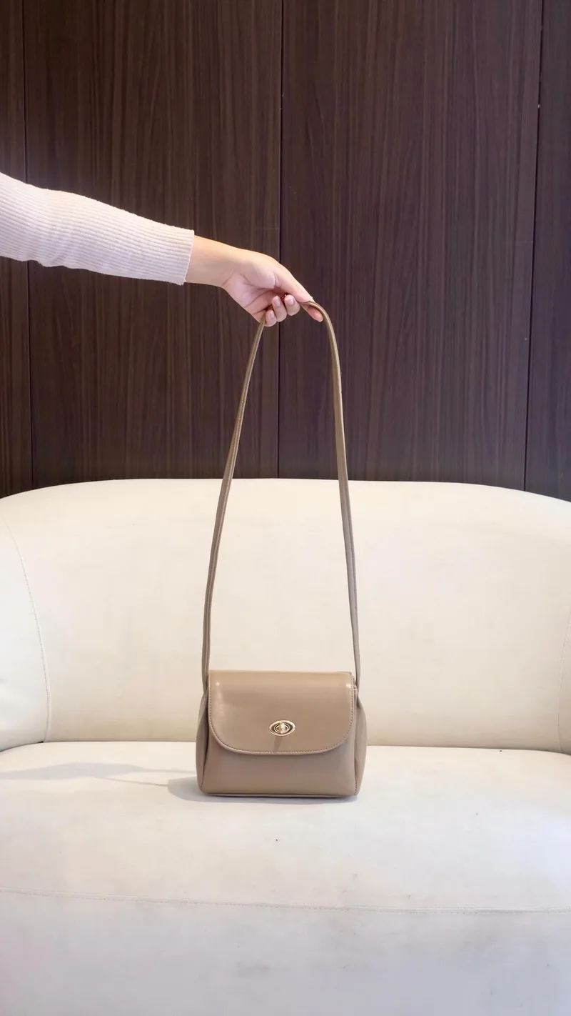 Tangan memegang tali panjang tas selempang kecil warna taupe dengan penutup lengkung dan kunci putar keemasan, di depan sofa putih berlatar dinding kayu gelap