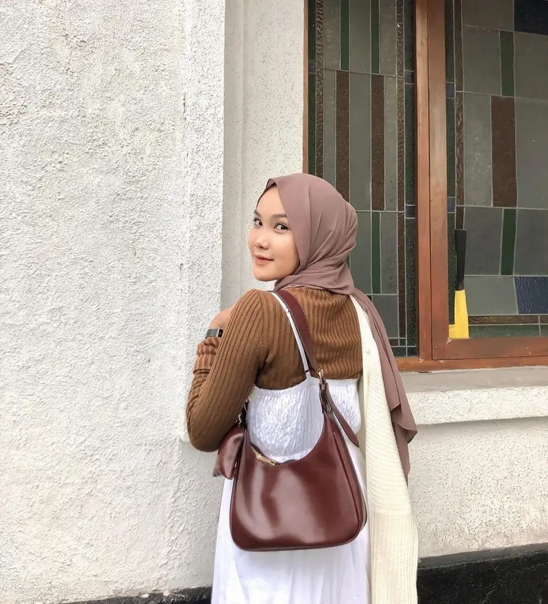 Perempuan berhijab cokelat susu, atasan rajut cokelat, dan gaun putih menyandang tas bahu cokelat kemerahan berbentuk bulan sabit, berdiri di depan dinding putih dan jendela kaca patri