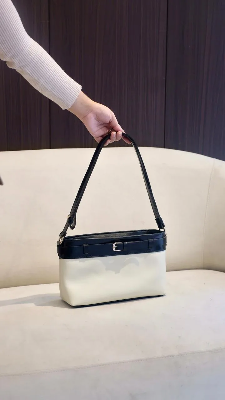 Tangan memegang tas bahu dua warna, bagian atas biru dongker dengan gesper dan badan putih gading, di depan sofa putih berlatar dinding kayu gelap