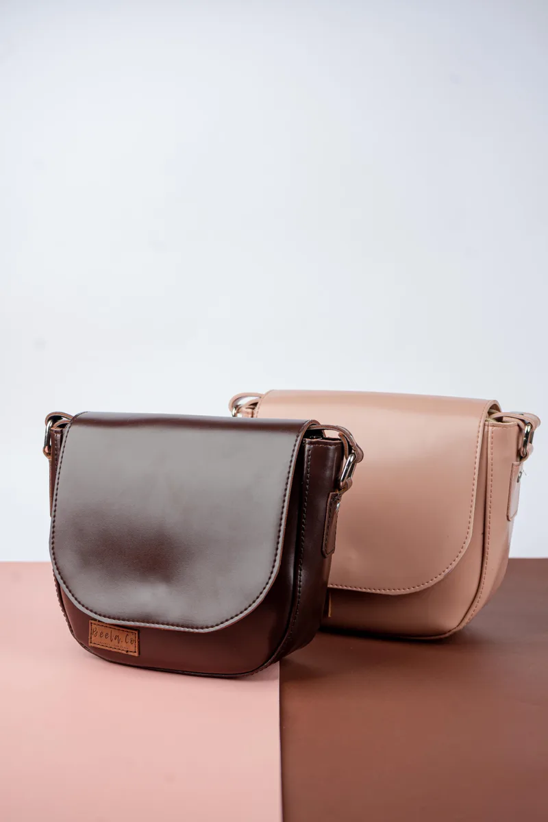 Dua tas selempang berflap setengah lingkaran berwarna cokelat tua dan merah muda nude dengan label kulit kecil di depan, berdiri di atas alas merah muda dan cokelat