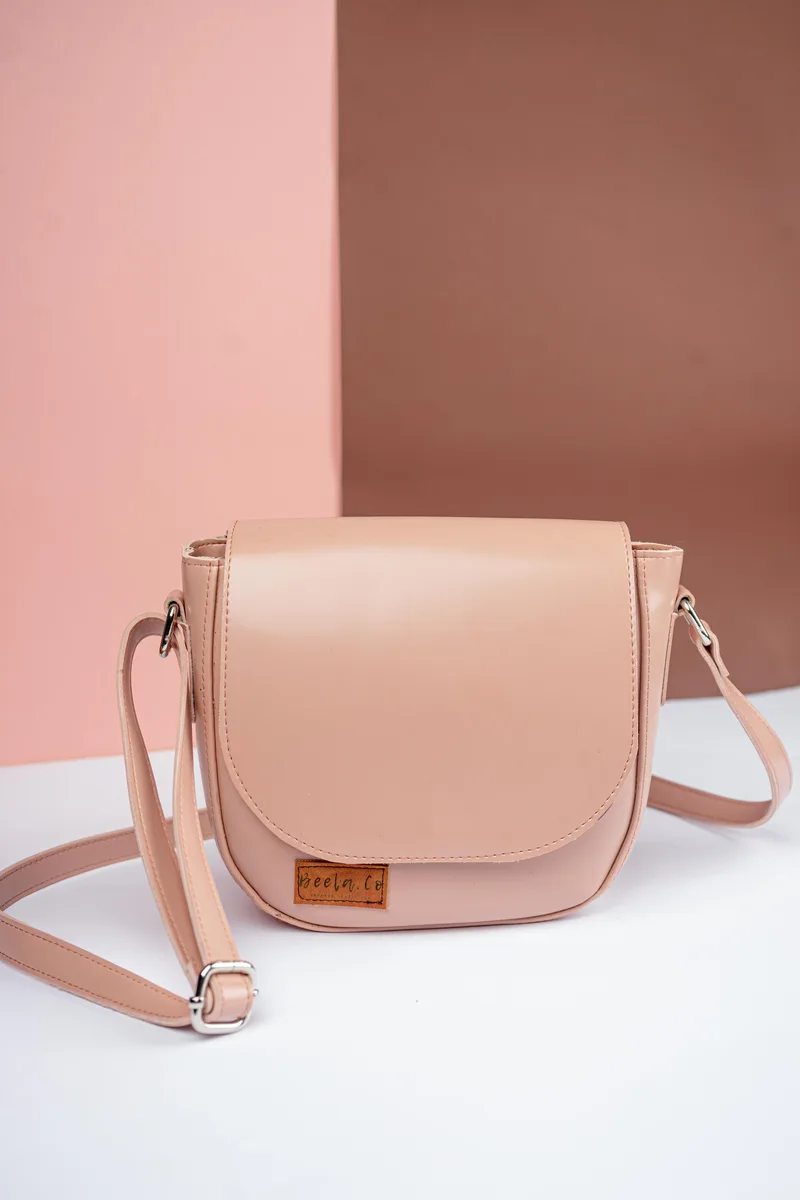Tas selempang berwarna merah muda nude dengan flap setengah lingkaran, label kulit kecil di bagian depan, dan tali panjang yang dapat disetel, berdiri di atas alas putih dengan latar merah muda dan cokelat