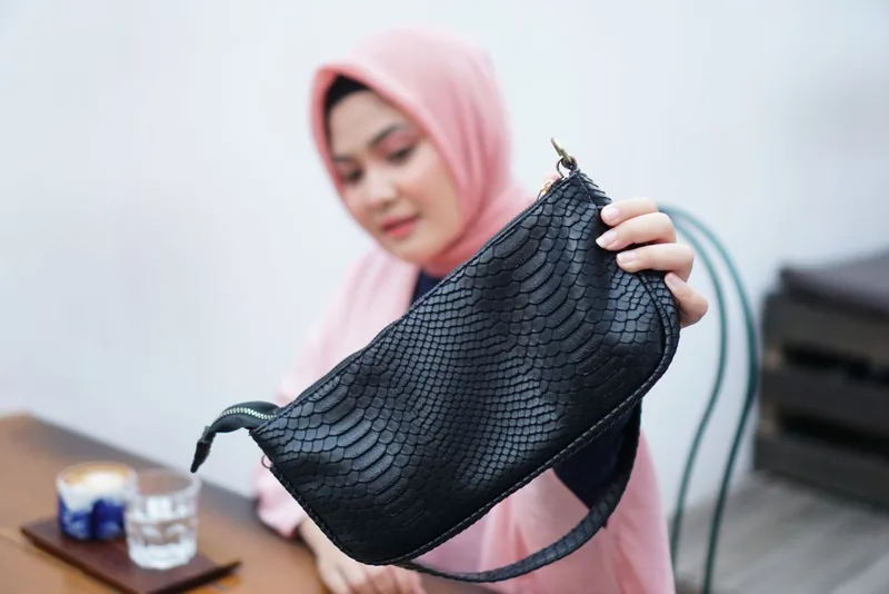 Seorang model berhijab merah muda memegang tas bahu hitam bertekstur timbul menyerupai sisik ular di depan tubuhnya, dengan meja berisi gelas di latar depan