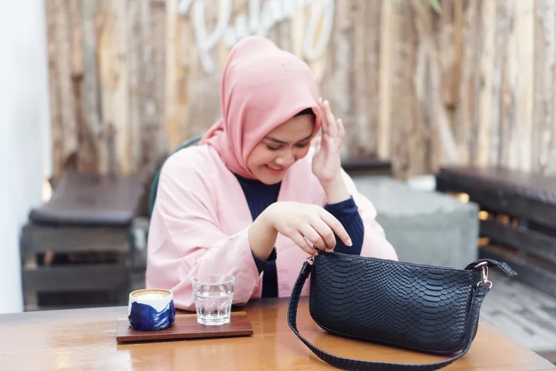 Seorang model berhijab merah muda tersenyum sambil membuka tas bahu hitam bertekstur di atas meja kayu, dengan gelas minuman di sampingnya