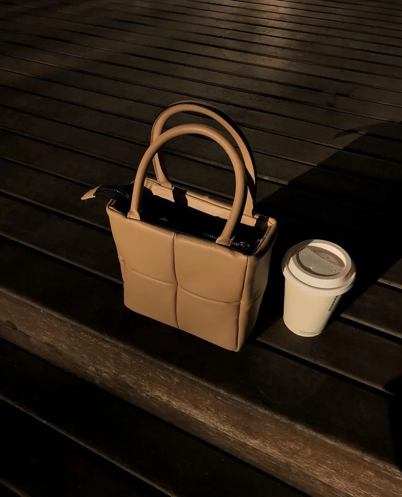 Tas jinjing warna cokelat muda berpanel kotak dengan dua pegangan bulat di atas meja kayu gelap, di sebelah gelas kopi kertas putih