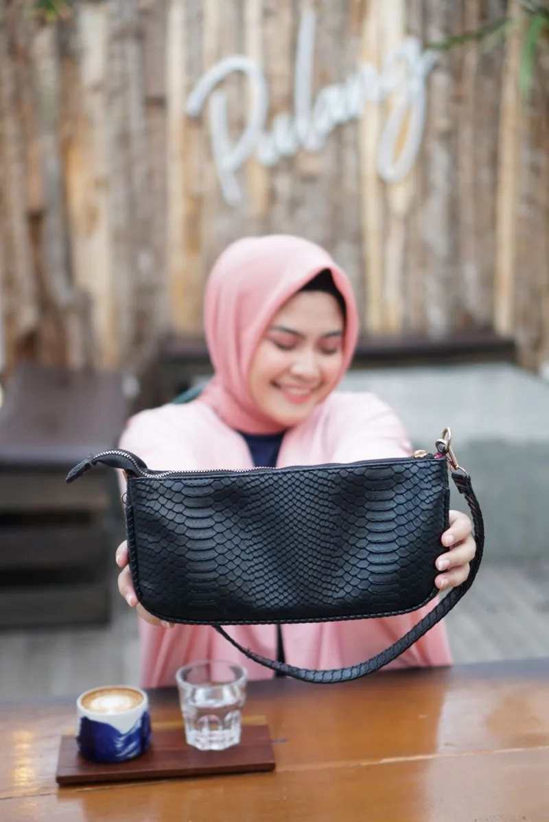Perempuan berhijab merah muda tersenyum sambil memegang tas bahu hitam bertekstur sisik di atas meja kafe dengan cangkir kopi dan gelas air