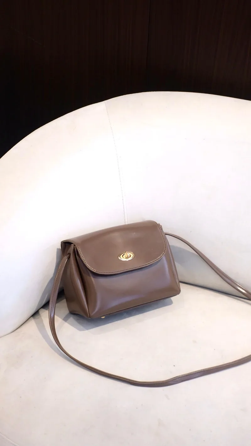 Tas selempang kecil berwarna cokelat dengan penutup flap dan kunci putar emas, tali panjang terjuntai di atas kursi berlapis kain putih