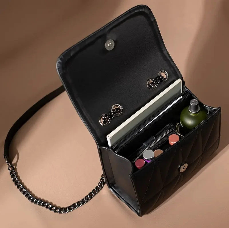 Tas hitam bermotif quilting dengan tutup terbuka memperlihatkan isi berupa buku catatan, botol parfum, lipstik, dan dompet ritsleting, dengan tali rantai gelap di samping