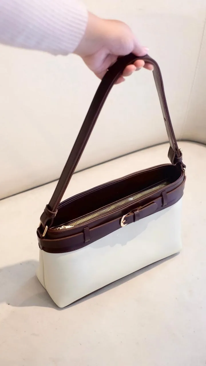 Tangan memegang tas bahu dua warna dengan badan putih gading dan bagian atas cokelat tua, ritsleting emas terbuka dan detail gesper kecil di depan