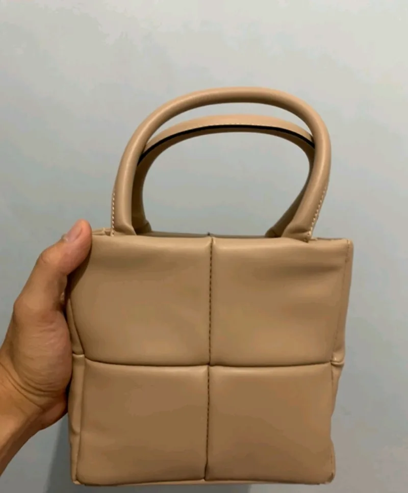 Tangan menggenggam tas tangan kecil warna nude dengan panel empuk berpola kotak dan dua pegangan bulat