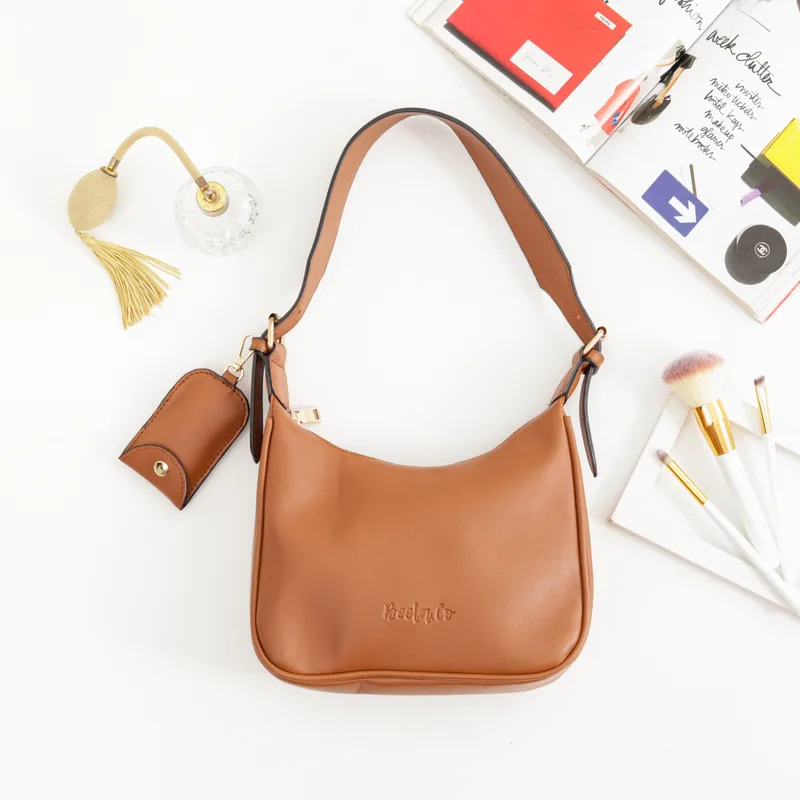 Tas bahu warna cokelat terang dengan logo Beela.Co timbul di bagian depan dan pouch mini di tali, ditata di atas meja putih bersama botol parfum dan kuas rias