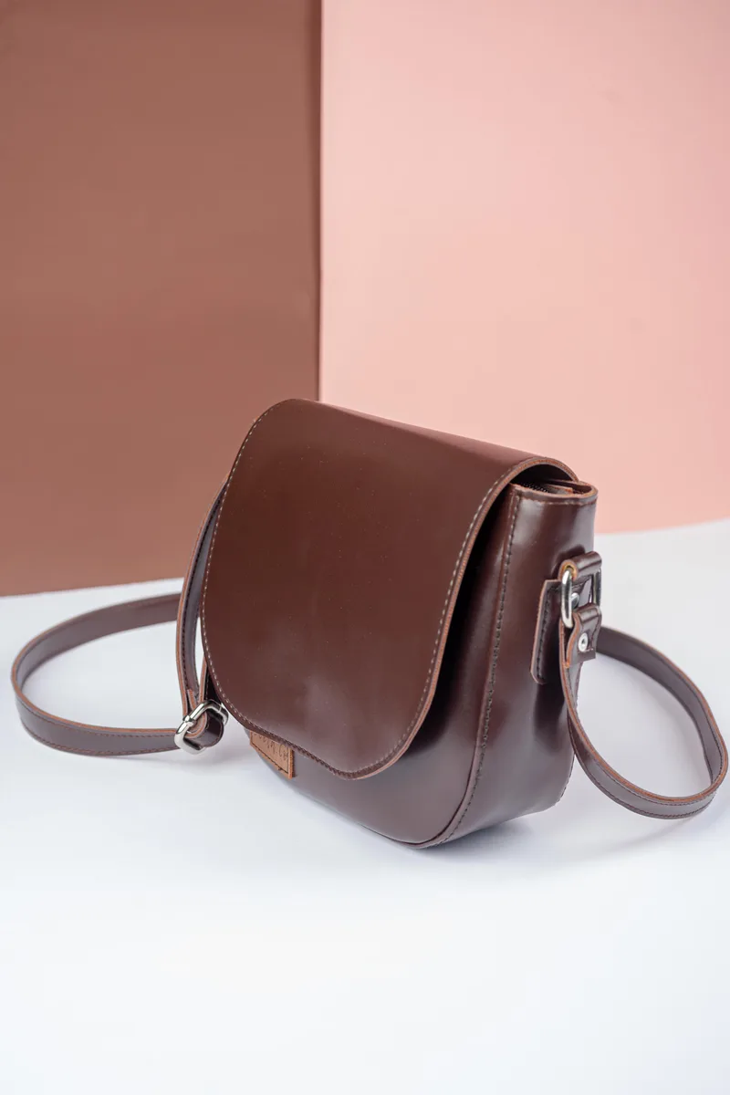 Tas selempang cokelat tua dengan tutup setengah lingkaran dan tali cokelat, difoto miring di atas alas putih dengan latar merah muda