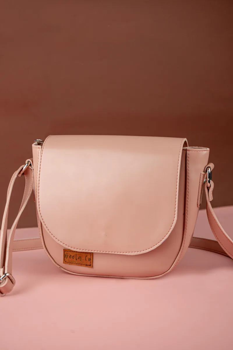 Tas selempang warna nude dengan tutup setengah lingkaran dan label kulit kecil bertuliskan Beela.Co di bagian depan, berdiri di atas alas merah muda