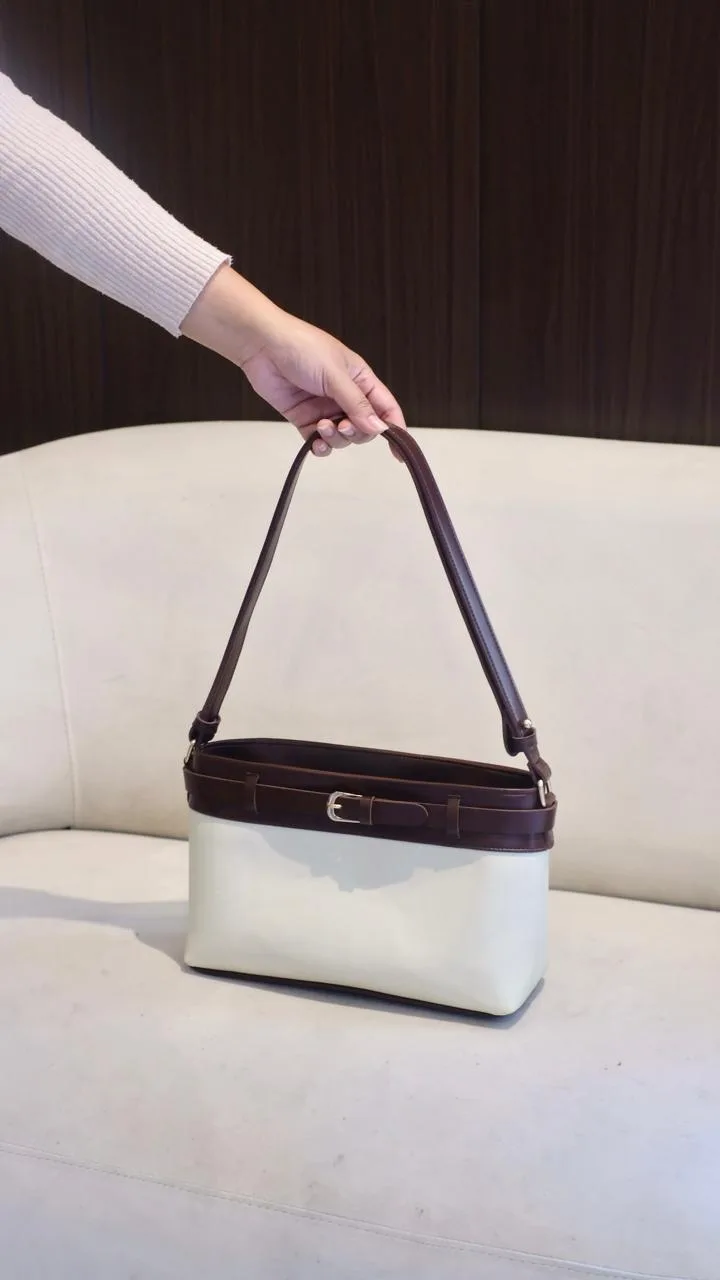 Tangan menjinjing tas bahu dua warna, bagian atas dan tali maroon dengan badan tas krem, di depan sofa putih