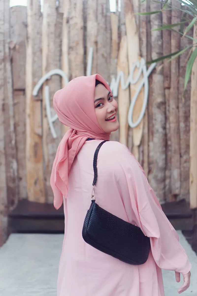 Perempuan berkerudung merah muda dan berbaju merah muda menoleh ke belakang sambil menyandang tas bahu hitam bertekstur sisik
