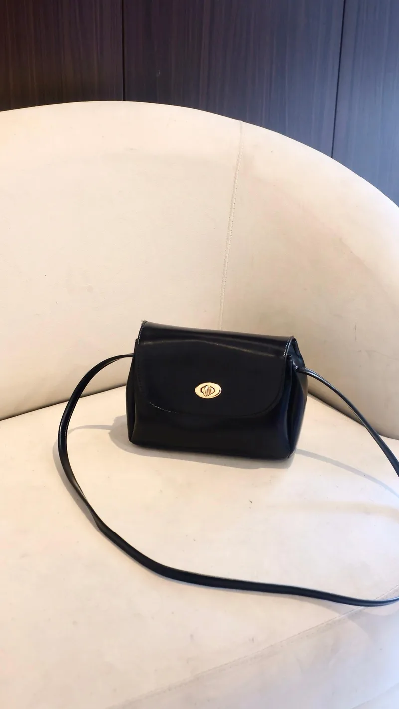 Tas selempang mungil hitam dengan flap dan kancing putar emas, tali hitam panjang melingkar di atas sofa putih