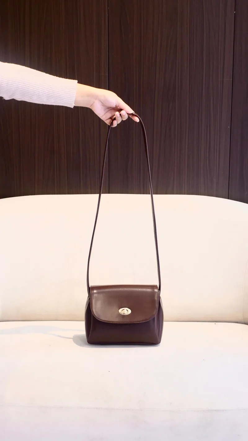 Tangan memegang ujung tali panjang tas selempang mungil warna maroon dengan kancing putar emas, tas menggantung di atas sofa putih
