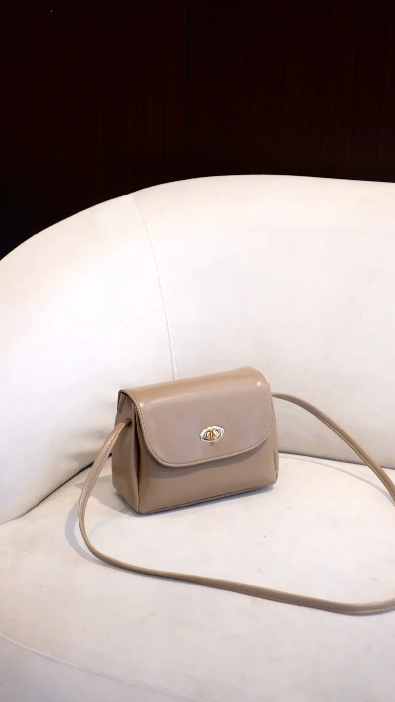 Tas selempang mungil warna taupe dengan kancing putar emas dan tali senada, diletakkan di atas sofa putih dengan latar gelap