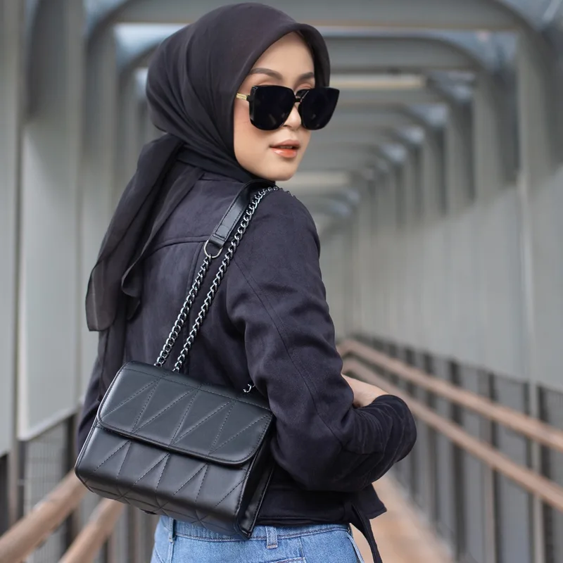 Perempuan berkerudung hitam dan berkacamata hitam menoleh ke kamera sambil menyandang tas selempang hitam bertali rantai di jembatan penyeberangan