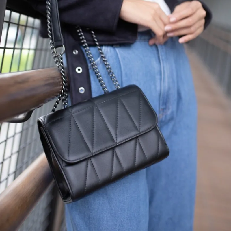 Tas selempang hitam bertekstur jahitan geometris dengan tali rantai hitam, dipakai di sisi pinggang oleh orang bercelana jeans biru