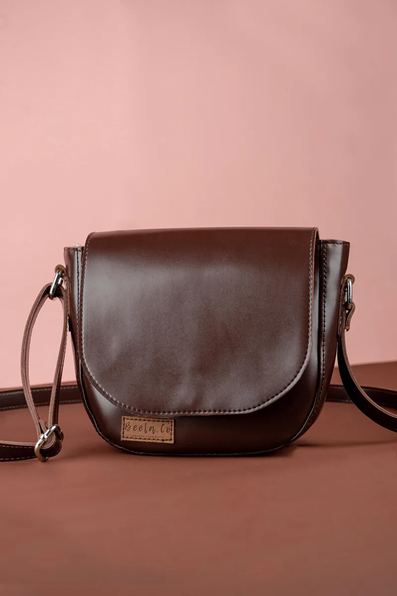 Tas selempang cokelat tua dengan tutup setengah lingkaran, tali kulit, dan label kecil Beela.co di bagian depan, berdiri di atas alas cokelat dengan latar merah muda