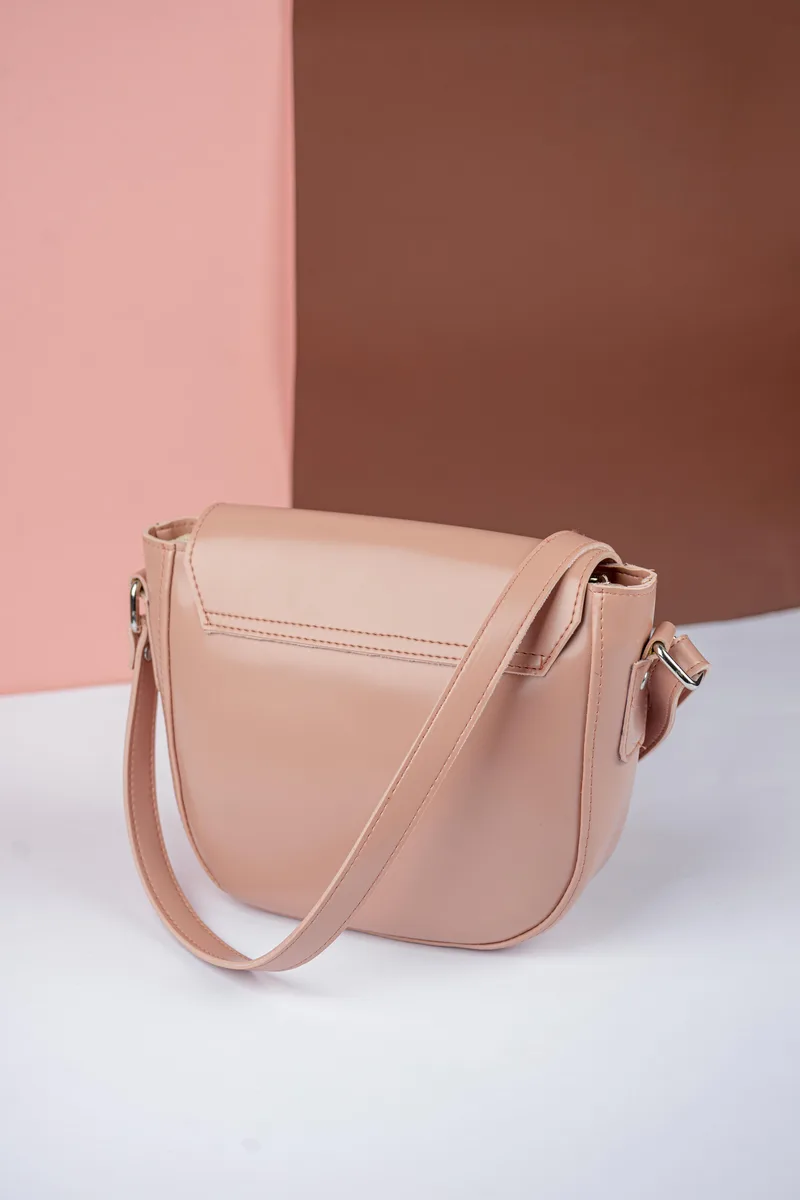 Tas selempang warna nude dengan tutup setengah lingkaran dilihat dari sisi belakang, tali kulit senada melengkung di atasnya, di atas alas putih dengan latar merah muda dan cokelat