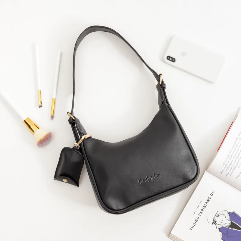 Tas bahu Benita berwarna hitam dengan tulisan Beela.Co timbul dan aksesori kecil berpengait, difoto dari atas di atas alas putih bersama kuas rias, ponsel, dan buku terbuka