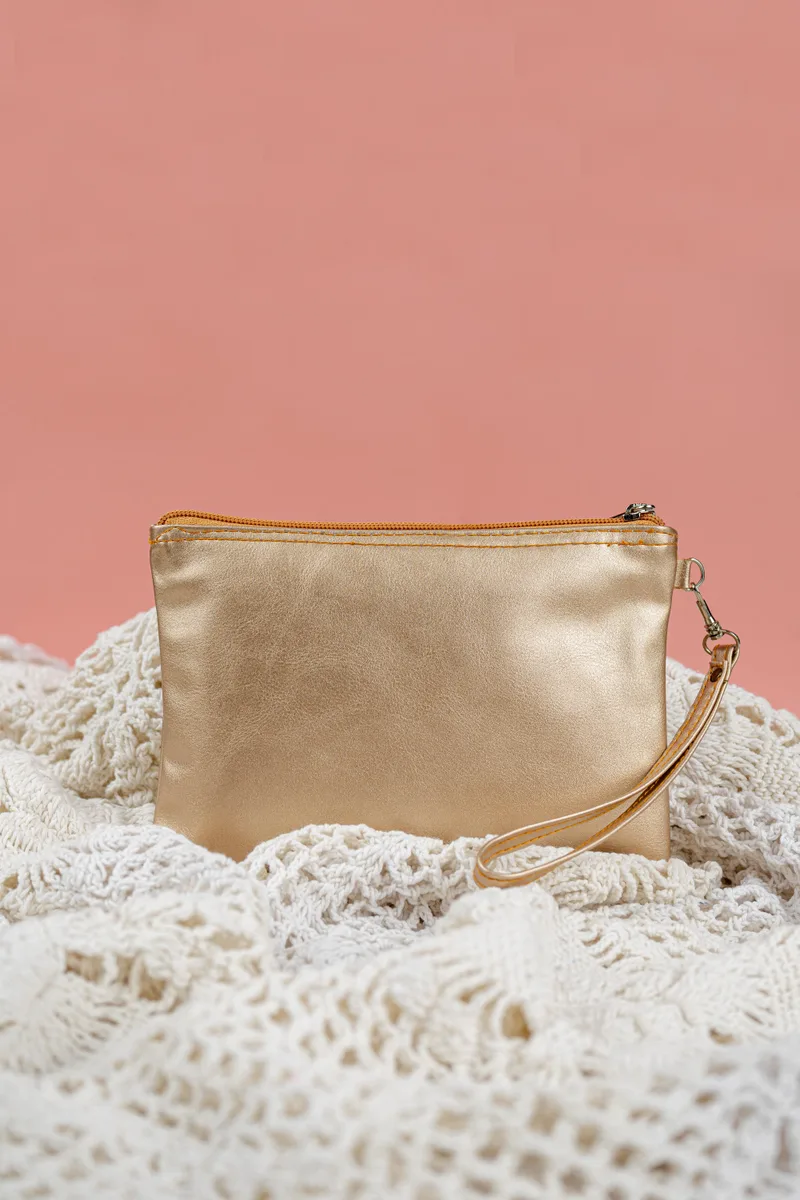Pouch berwarna emas metalik dengan ritsleting di bagian atas dan tali tangan berpengait, diletakkan di atas kain rajut putih dengan latar dinding merah muda
