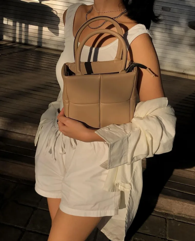 Perempuan berbaju putih memeluk tas berpanel kotak warna cokelat muda di depan dada, dengan kemeja krem tersampir di lengan