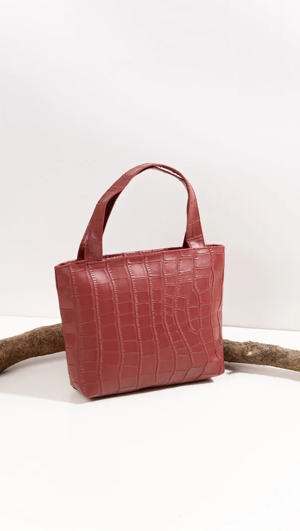 Tas jinjing kecil bertekstur croco warna merah bata dengan dua pegangan, diletakkan di atas latar putih dengan sepotong kayu di belakangnya