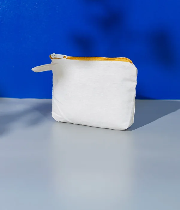 Pouch kain berwarna putih polos dengan ritsleting kuning di bagian atas dan tali kecil di sisi kiri, berdiri di atas alas abu-abu dengan latar biru
