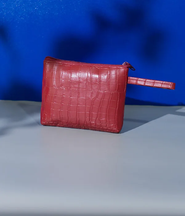 Pouch berwarna merah dengan tekstur kotak menyerupai kulit buaya, ritsleting di bagian atas, dan tali kecil di sisi kanan, berdiri di atas alas abu-abu dengan latar biru