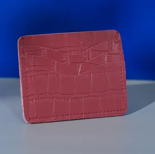 Card holder berwarna merah bata dengan tekstur kulit buaya timbul dan slot kartu bertingkat, berdiri di atas permukaan abu-abu dengan latar biru