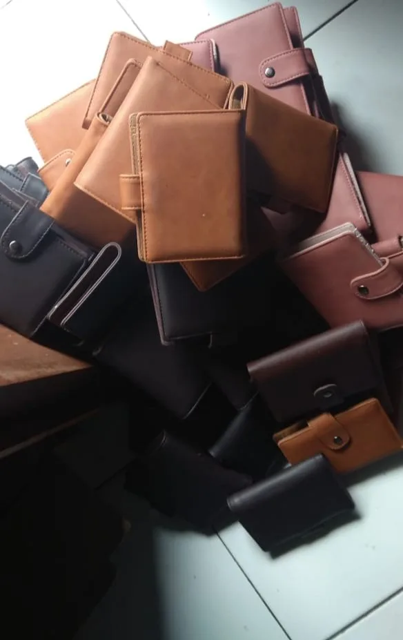 Tumpukan dompet lipat berbagai warna cokelat muda, cokelat tua, dan hitam, sebagian dengan tali kancing, menumpuk di atas meja