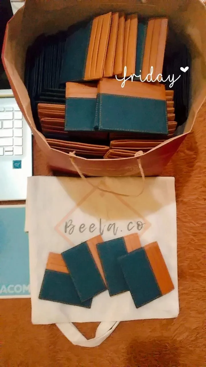 Card holder dua warna hijau tua dan oranye di dalam tas kertas dan di atas tas kain bertuliskan Beela.co