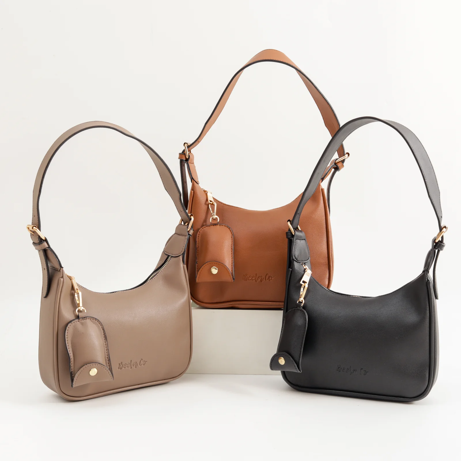 Tiga Benita Bag Beela.Co warna taupe, tan, dan hitam, masing-masing dengan pouch kecil, berdiri di pedestal putih